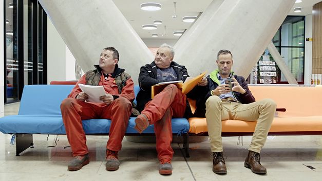 Still from the film "For the Many – The Vienna Chamber of Labour" by Constantin Wulff: In the lobby of an office building, two men in work wear and one man in office attire are sitting on a bench, looking at something off camera.