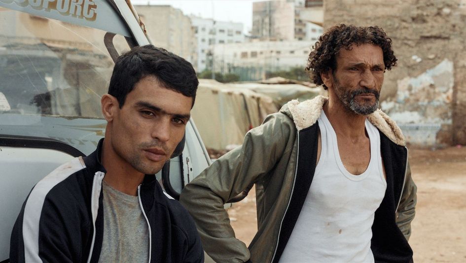 Filmstill aus HOUNDS: Zwei Männer in einfacher Kleidung stehen in einer Stadt neben einem Auto.