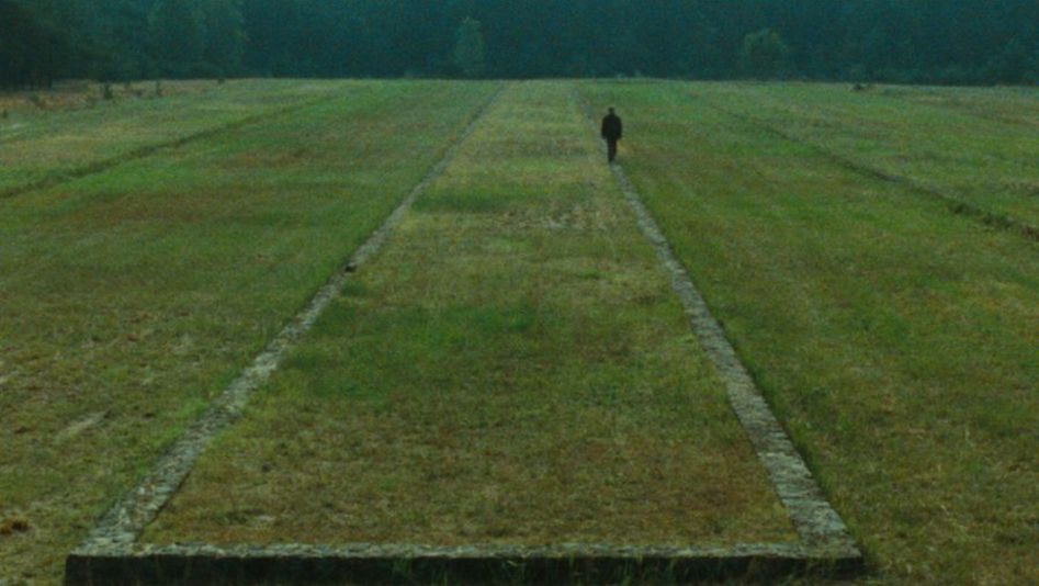 Film still from SHOAH: You can see a landscape with a meadow and forest in the background. The remains of a building can be seen in the meadow, a person is walking along it.