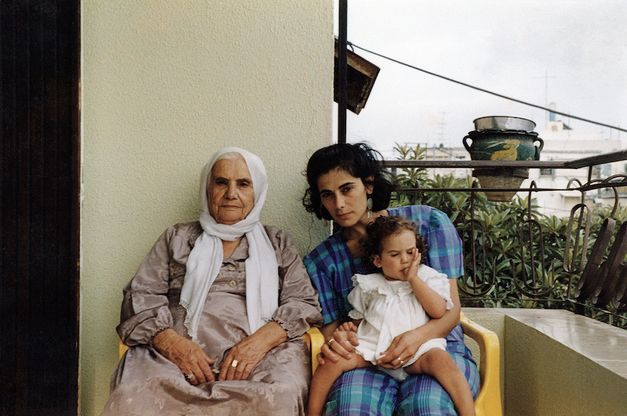 Film still from BYE BYE TIBERIAS: An old and a young woman are sitting next to each other on a balcony. The younger woman has a little girl on her lap.