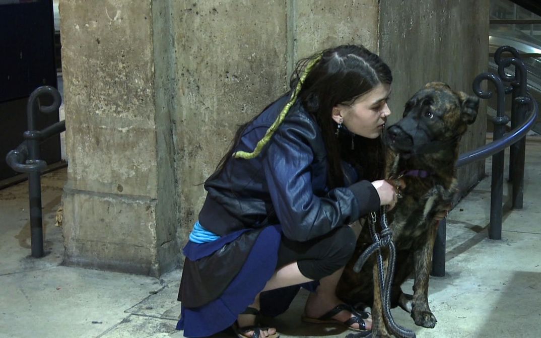Film still from GEOGRAPHIE HUMAINE: A young woman crouches next to a dog on a street corner.