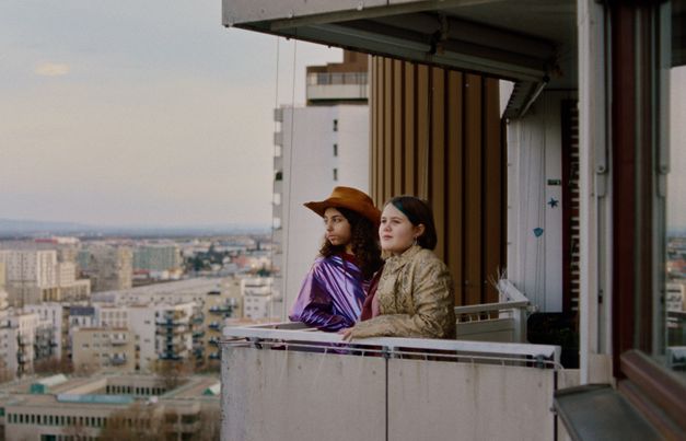 Film still from WENN DU ANGST HAST NIMMST DU DEIN DEIN HERZ IN DEN MUND UND LÄCHELST by Marie-Luise Lehner. Two young women stand on the balcony of a high-rise building and look into the distance. 