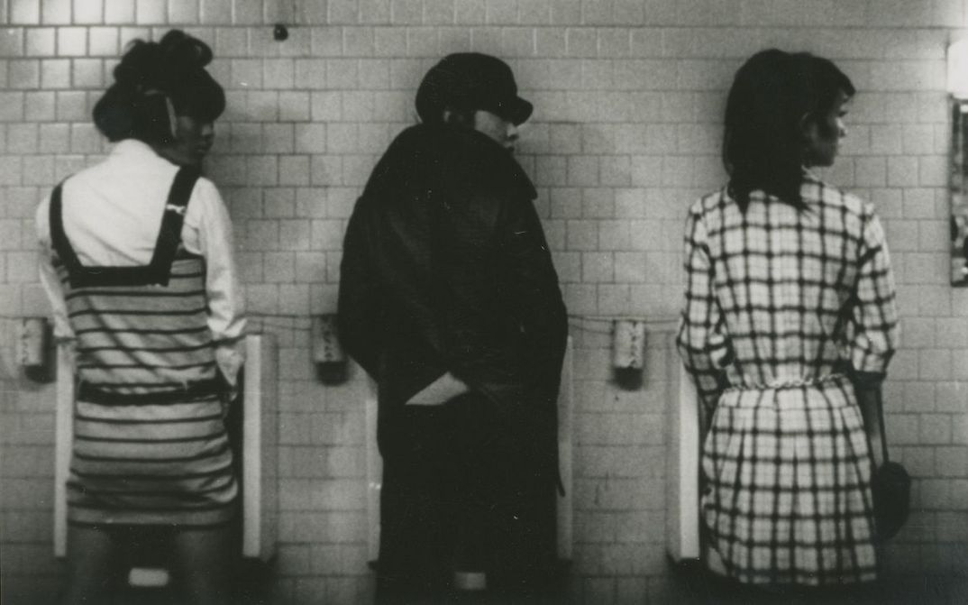Film still from FUNERAL PARADE OF ROSES: Three people in extravagant dresses in front of three urinals, all looking to the right.