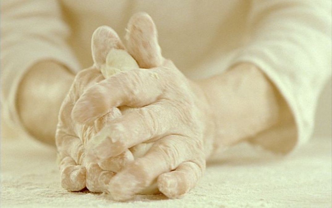 Filmstill aus CUM PANE. Großaufnahme von zwei Händen, die Teig kneten.