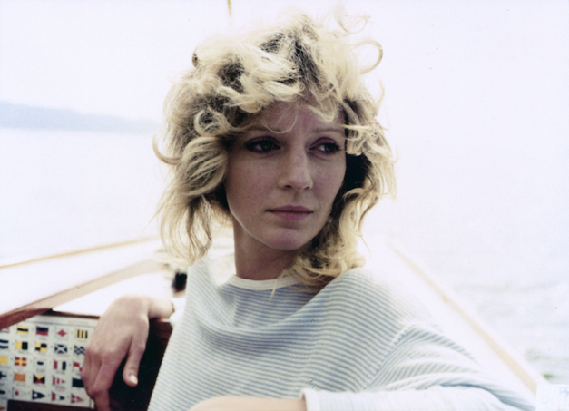 Film still from DIE ALLEINSEGLERIN: A young woman with curly blonde hair stands at the bow of a sailing boat.