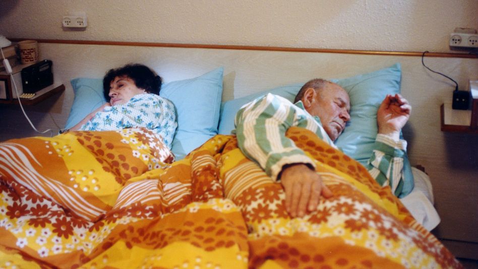 Filmstill from HABEHIRA VEHAGORAL: A woman and a man lie asleep in bed. The bed linen and pyjamas are patterned and colourful.