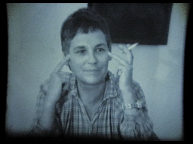 A woman sits at a table, points to her temples with her fingers and holds a cigarette.