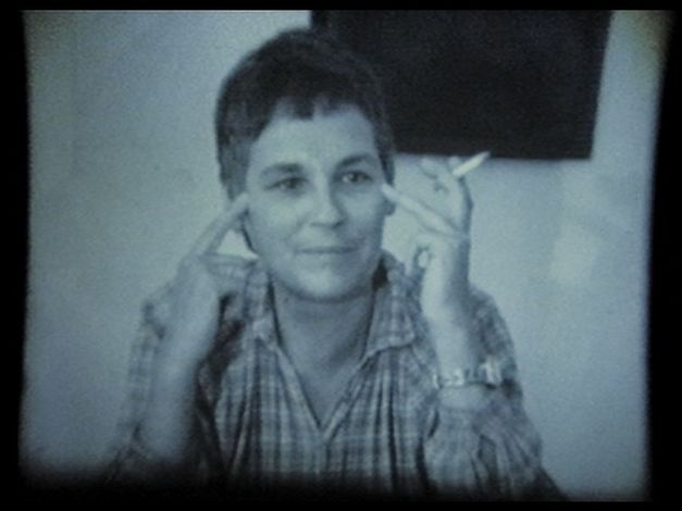 Frau sitzt an einem Tisch, zeigt mit den Fingern auf ihre Schläfen und hält eine Zigarette.