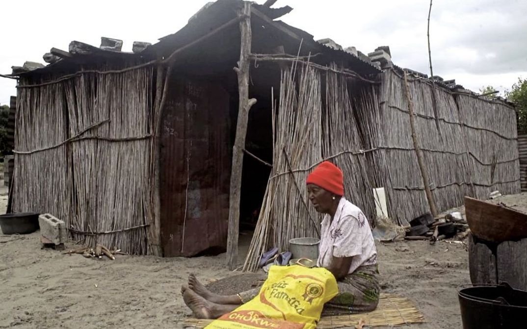 Filmstill aus "The Nights Still Smell of Gunpowder" von Inadelso Cossa. Zu sehen ist eine Frau mit roter Mütze sitzt vor einer Hütte aus Stöckern auf dem Boden.