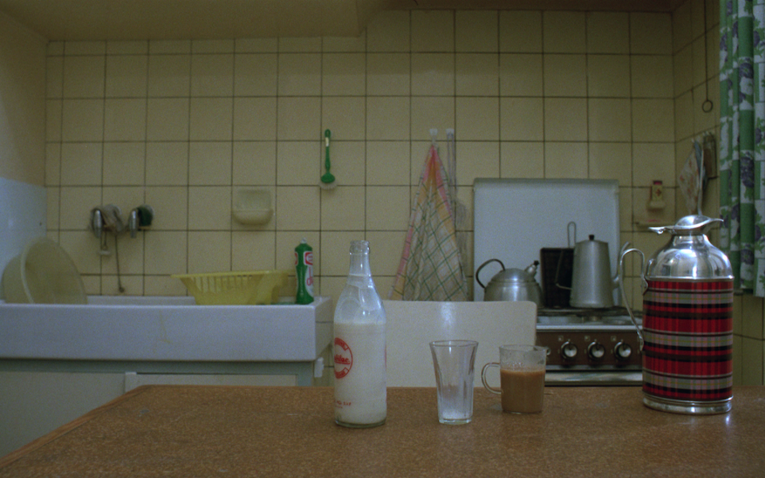 Film still from JEANNE DIELMAN: You can see a kitchen, in the background sink and stove, in the foreground a table on which there is a milk bottle and glasses.