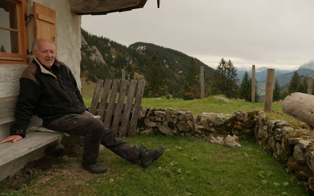 Film still from WERNER HERZOG – RADICAL DREAMER: Werner Herzog sits on the bench in front of a mountain hut.