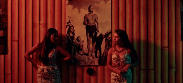 Two women stand in front of a wall of bamboo poles on which hangs a picture of Johnny Weissmuller as Tarzan.