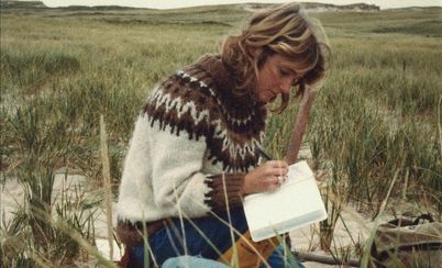 Still from "Geographies of Solitude" by Jacquelyn Mills. The protagonist Zoe Lucas is sitting in a landscape of dunes, writing in a notebook