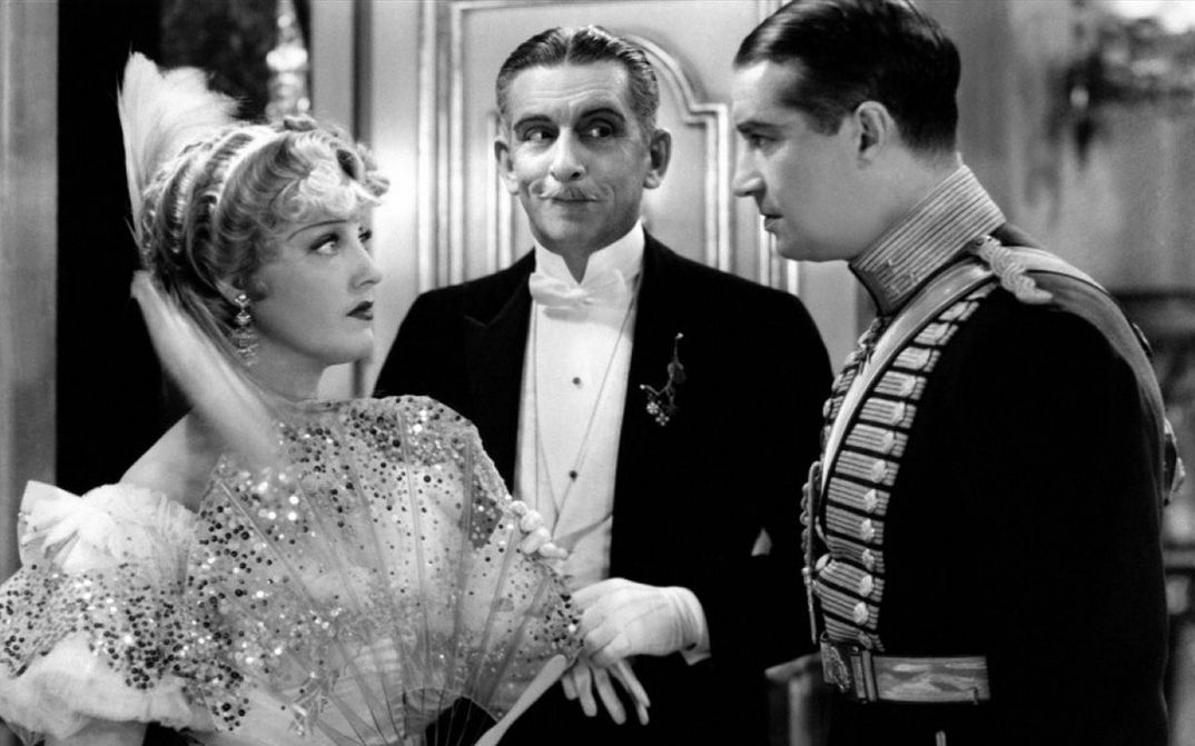 Film still from THE MERRY WIDOW: A young woman and a young man, both festively dressed, look at each other. An elderly gentleman stands in the background.