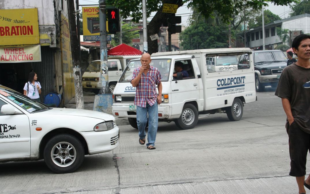 Film still from MANILA: A man crosses a road on which several cars are driving. He has a cigarette in his mouth.