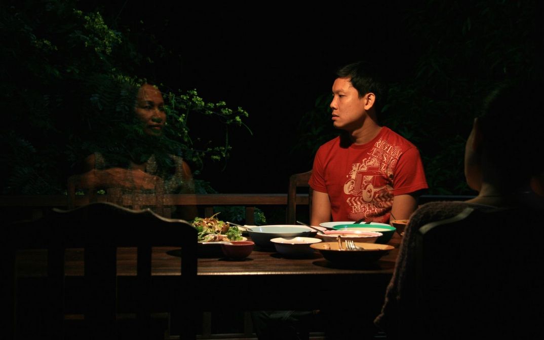 Filmstill aus UNCLE BOONMEE ERINNERT SICH AN SEINE FRÜHEREN LEBEN: Ein Mann sitzt in der nächtlichen Dunkelheit an einem Tisch und blickt auf eine durchscheinende Frau.