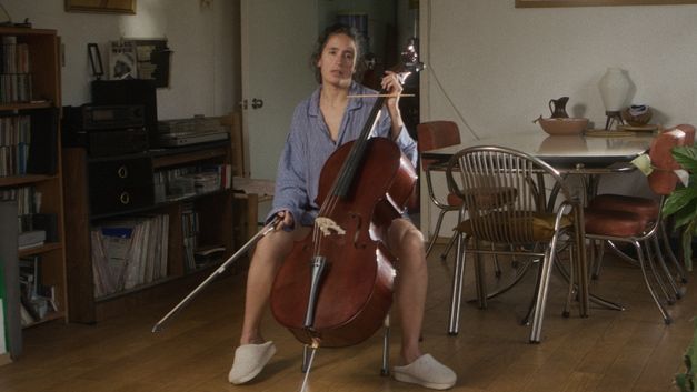 Person playing cello in living room surrounded by furniture and plants.