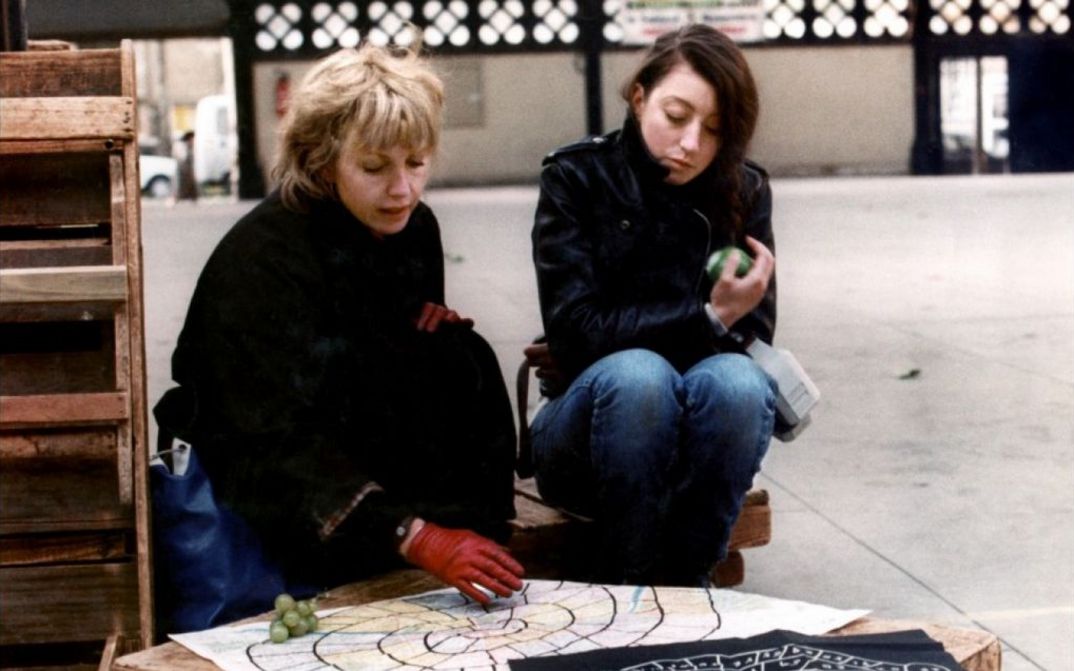 Film still from LE PONT DU NORD: Two women are sitting next to each other outdoors. They are looking at a cloth lying on a low table.