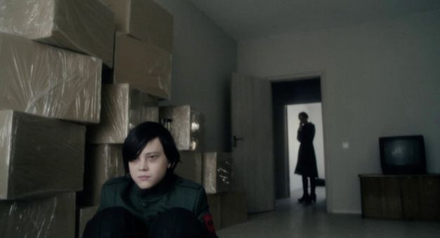 Film still from CADET by Adilkhan Yerzhanov. The image shows a boy in uniform crouching on the floor and the silhouette of a person in the background in the door frame.