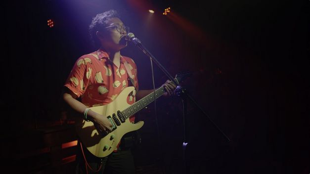 Filmstill aus QUEER AS PUNK von Yihwen Chen. Das Bild zeigt eine performende Person mit einer Gitarre und einem Mikrofon im Scheinwerferlicht.