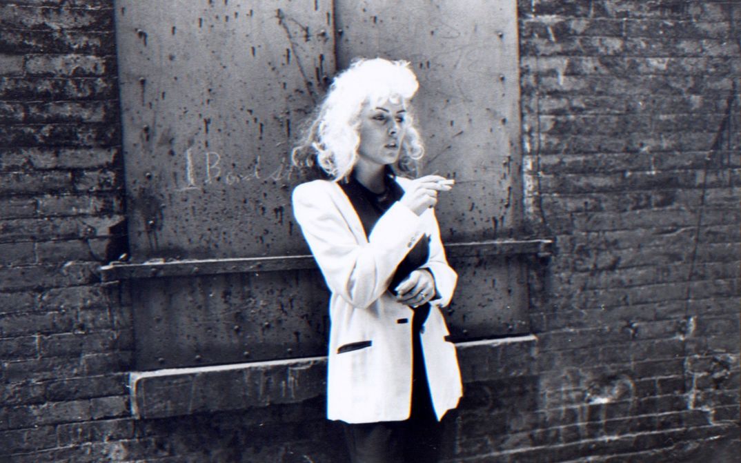 Filmstill from FOREIGNER: Eine Faru mit blonden Haaren und weißem Jackett lehnt rauchend an einer Backsteinwand und blickt nachdenklich nach rechts.