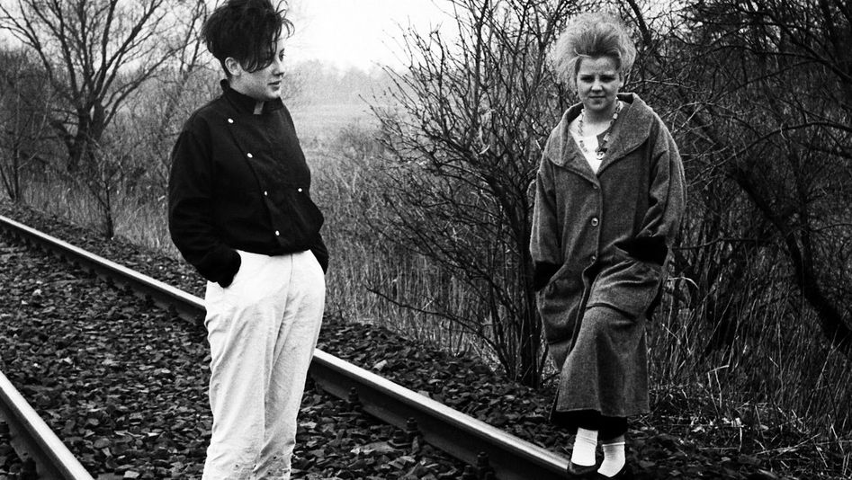 Filmstill aus WINTER ADÉ: Zwei Frauen stehen auf Bahnschienen. Im Hintergrund ist ein Feld und Bäume. Es wirkt trist und grau.
