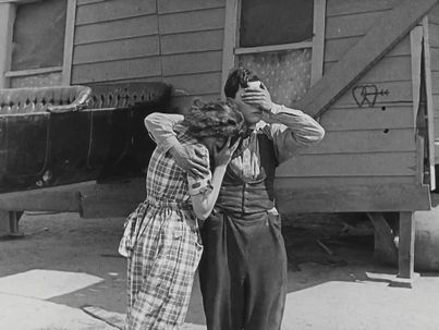 Filmstill aus ONE WEEK: Ein Mann und eine Frau stehen vor einem einfachen Holzhaus. Er hat den Arm um sie gelegt, beide halten ihre Hände vor die Augen.