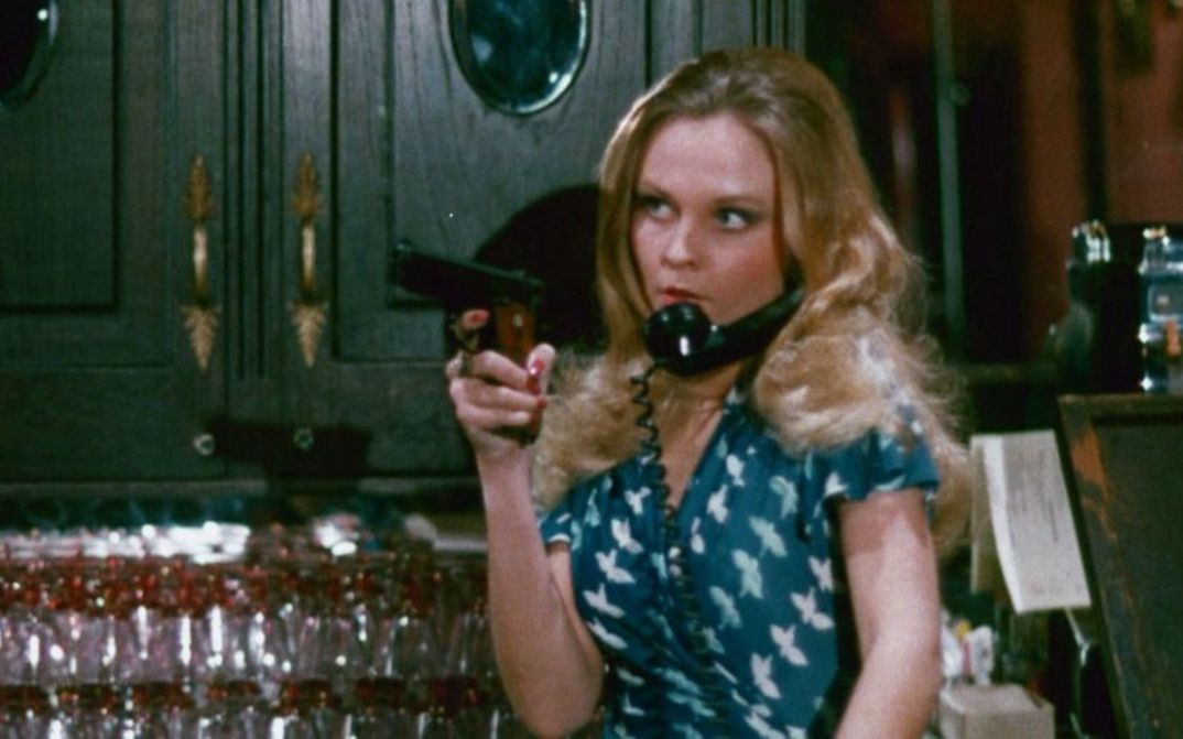 Film still from THE WORKING GIRLS: A woman is standing behind a bar, holding a gun in her hand. A telephone receiver is wedged between her head and shoulder.