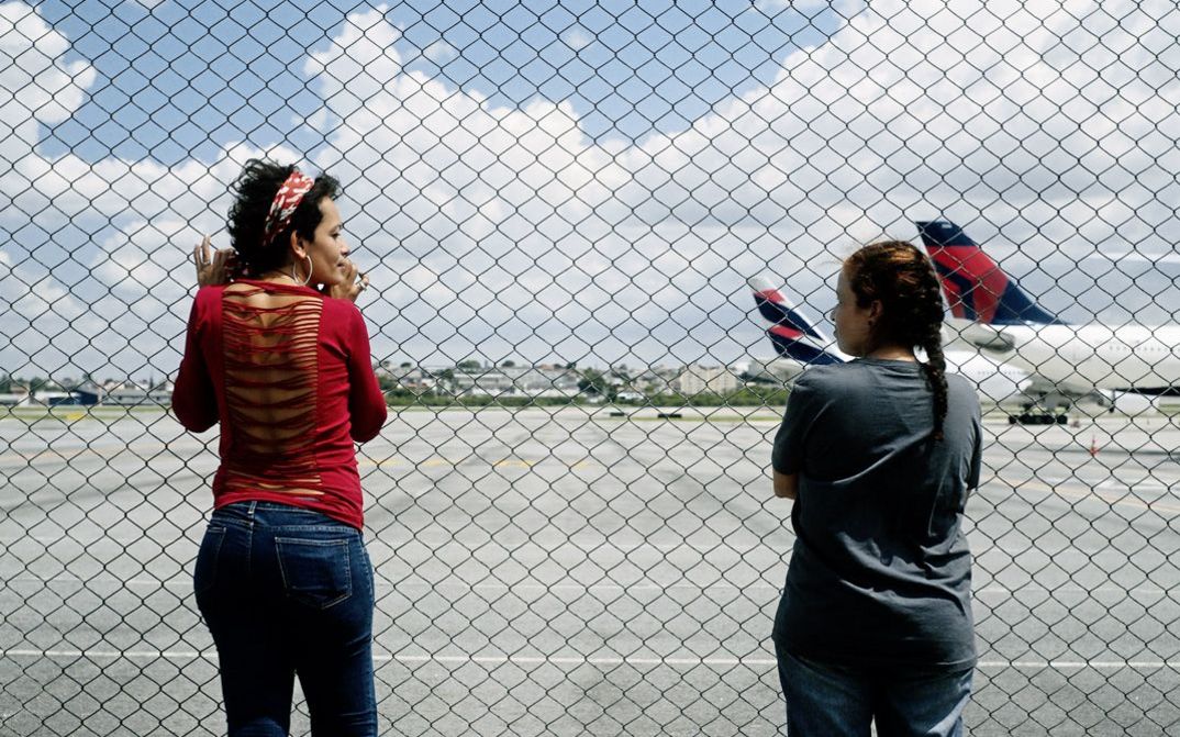 Still from the film "O Estranho" by Flora Dias and Juruna Mallon. Two women stand next to each other in front of a fence and look at each other. Two airplanes standing on an airport runway can be seen in the background.