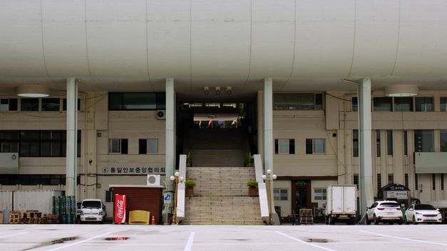 Film still from THE SENSE OF VIOLENCE by Mooyung Kim. The picture shows the ground floor of a large building. Cars are parked to the left and right of a staircase. 