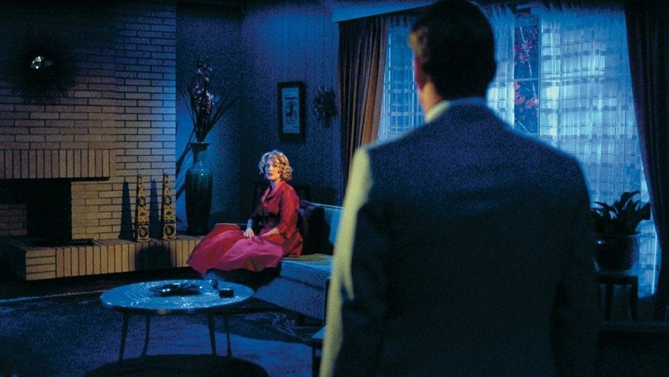 Filmstill from FAR FROM HEAVEN: A woman in a red dress is sitting in a living room. It is dark. A man can be seen from behind, looking in her direction.