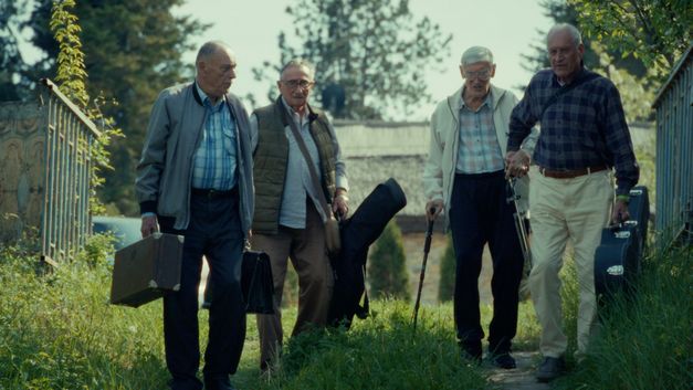 Film still from RESTITUCIJA, ILI, SAN I JAVA STARE Gare by Želimir Žilnik. The picture shows four older men with suitcases and instrument bags outdoors.