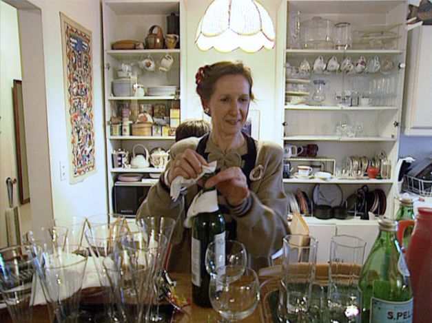 Filmstill from MANHATTAN STORIES: A woman in an apron wipes a wine bottle. Around her are many glasses and kitchen utensils.