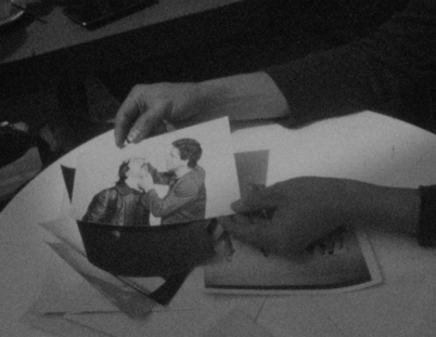 Still from the film „This Makes Me Want to Predict the Past“: A black and white photo in which two hands are holding another photo into the camera. On the photo, two men are pictured