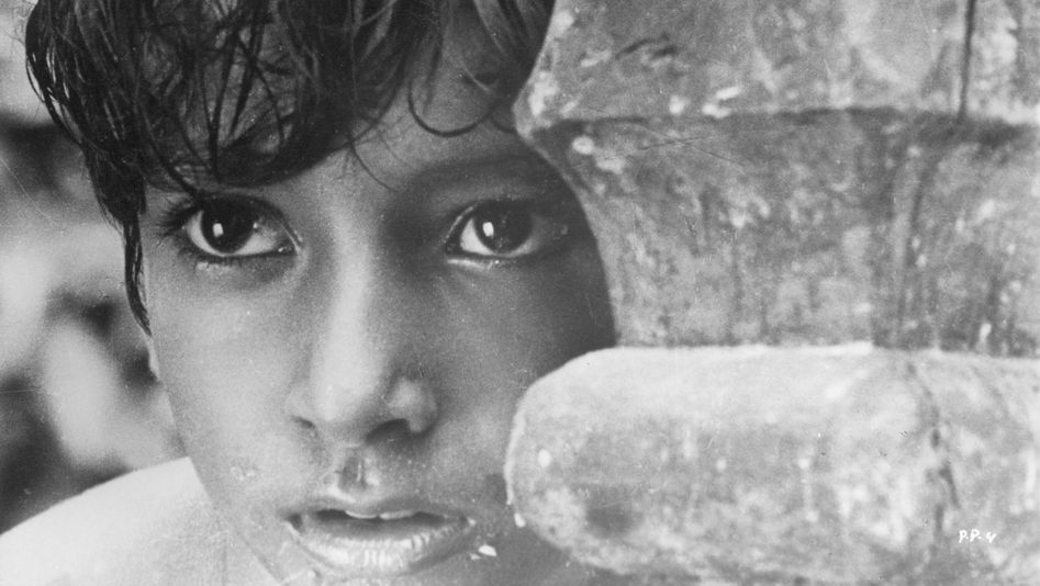 Filmstill from PATHER PANCHALI: A boy looks as if he is watching something and hiding behind a pillar or wood.
