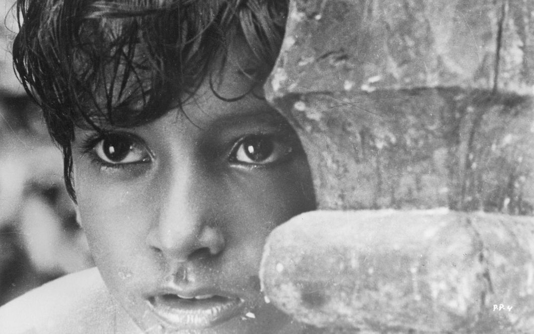 Filmstill from PATHER PANCHALI: A boy looks as if he is watching something and hiding behind a pillar or wood.
