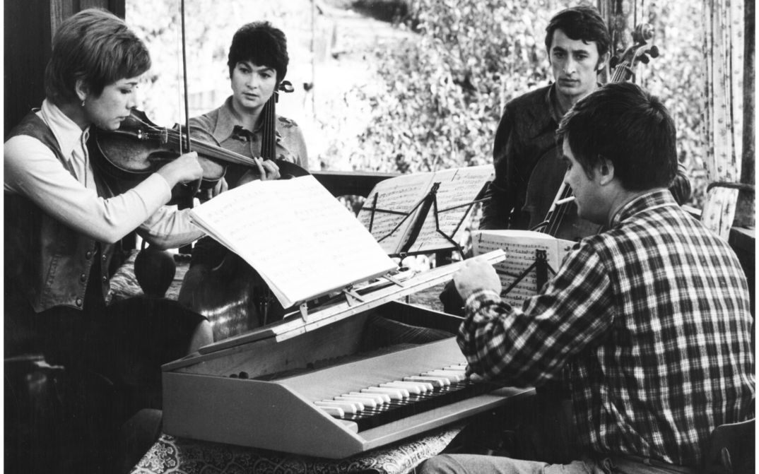Film still from PASTORALE: A group of musicians stand around a piano.