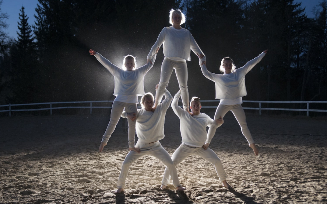 Film still from STUDIES ON THE ECOLOGY OF DRAMA: Five people, all dressed in white, form a small human pyramid. They stand in the dark on a sandy area and are illuminated by a spotlight.