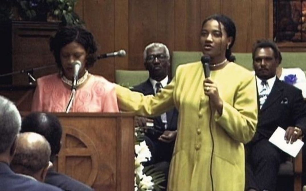 Film still from SUD: Two women are standing at a pulpit with microphones, with other people behind and in front of them.