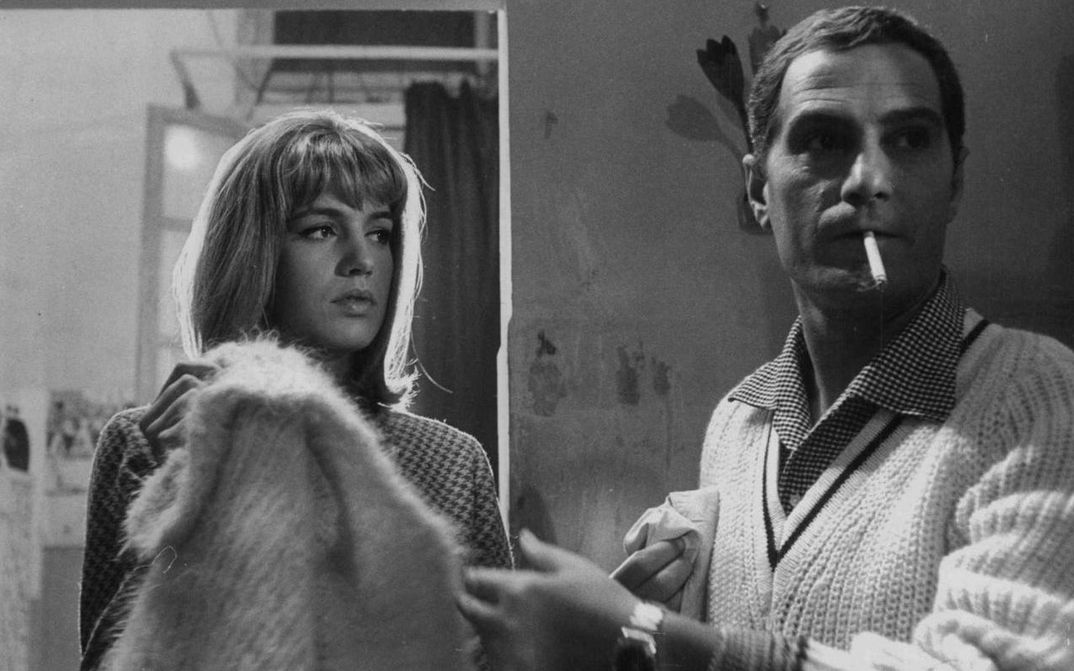 Film still from LA PARMIGIANA: A young woman and a man in a room. She looks at him, he is slightly turned away from her and has a cigarette in his mouth.