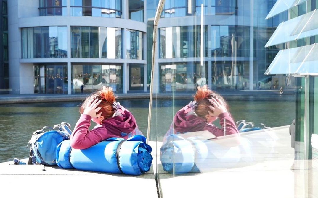 Filmstill aus AUFBRUCH: Eine junge Person sitzt am an einem Fluss, durch eine Spiegelung ist sie doppelt zu sehen. Auch am anderen Flussufer ist ein modernes Gebäude mit Glasfassade.