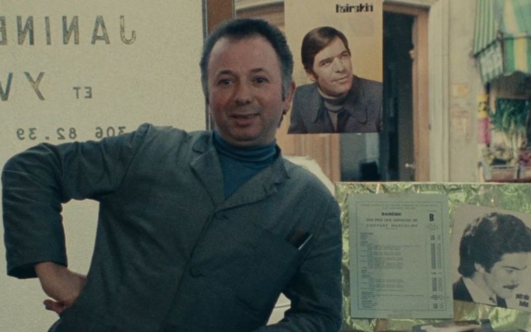 Film still from DAGUERRÉOTYPES: A man in a work coat with his hand on his hip. Behind him, photos of men and a vegetable stand.