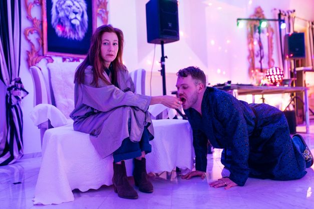 A woman sits in an armchair while a man kneels on all fours and eats from her hand in a colorfully lit room.