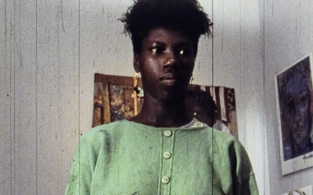 Still from the film „A Lover & Killer of Colour“ by Wanjiru Kinyanjui. Close-up of a woman. In the background, pictures hang on the wall.