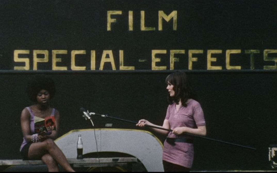 Film still from DARK SPRING. A woman holds a microphone out to another who is reading from a book. In the background the lettering Film Special Effects.