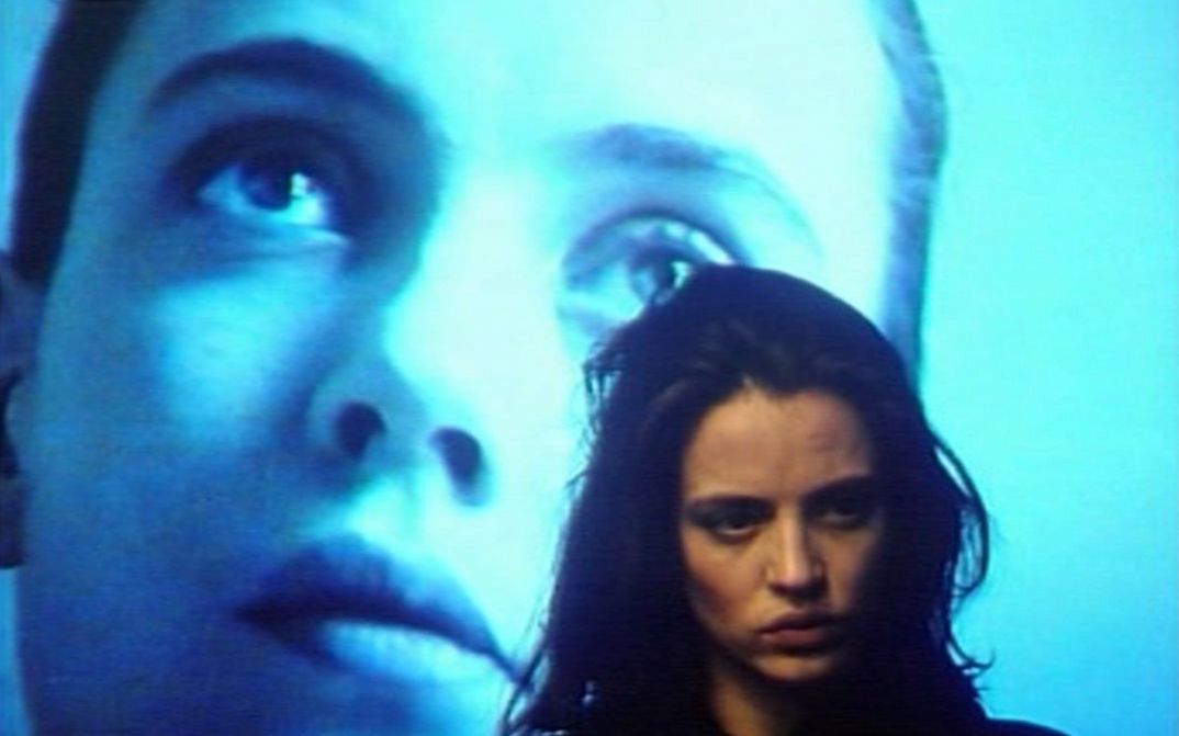 Film still from GINEVRA. Close-up of a woman, in the background a face is projected on a screen.