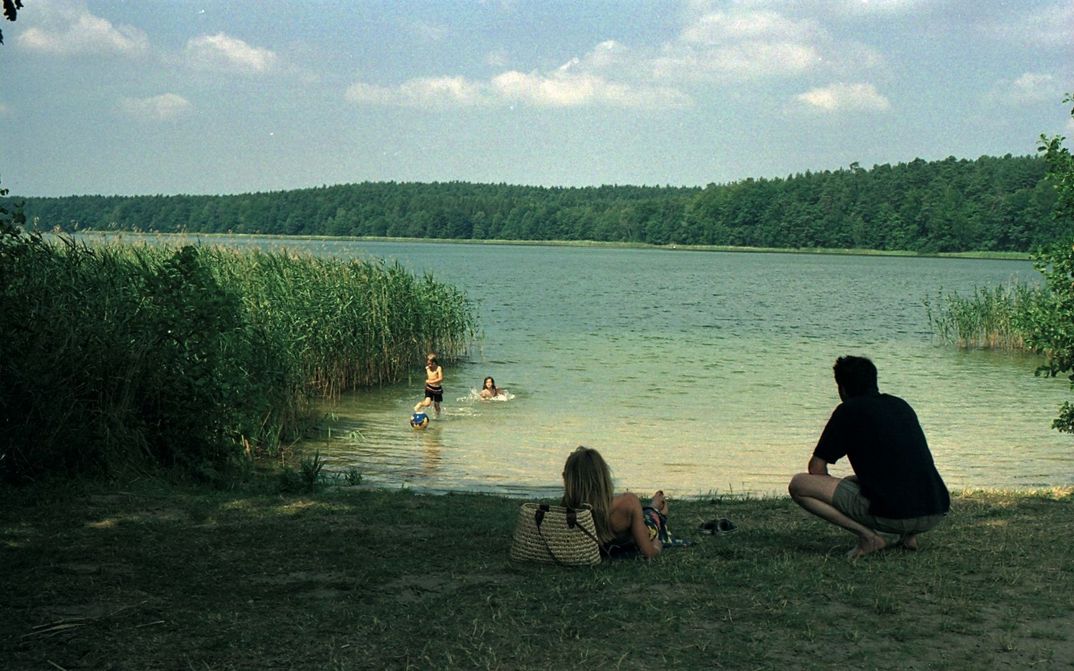 Filmstill aus FERIEN: Kinder im Wasser eines Sees, ein Mann und eine Frau liegen auf der Wiese davor. Die Sonne scheint, um den See ist Schilf und Wald.