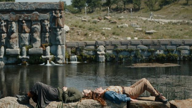 Two people lie side by side on a pier by the water, with a stone embankment in the background.
