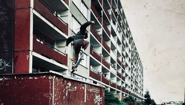 Person springt mit Skateboard vor langem sozialistischen Plattenbau.