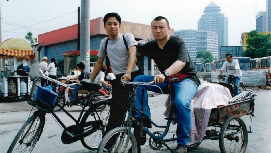 Film still from THERE'S A STRONG WIND IN BEIJING: Two young men on the street with their bicycles. In the background a city, buses and a street stall.